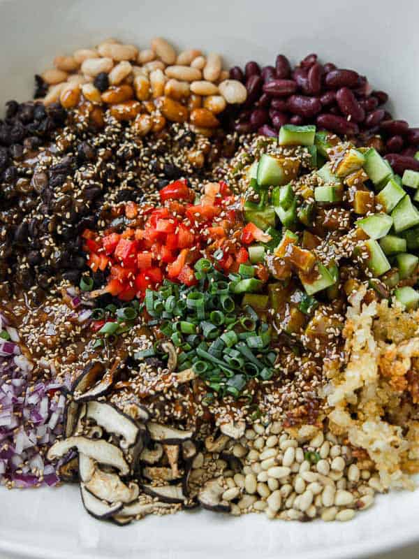 A colorful bowl with neatly arranged ingredients, including beans, diced cucumbers, red peppers, green onions, mushrooms, sesame seeds, pine nuts, chopped red onions, and crispy rice.